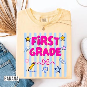 May include: A pale yellow t-shirt featuring the words "FIRST GRADE" in pink, with stars, paperclips, a pencil, and flowers. The shirt has a crew neck and short sleeves. The word "BANANA" is printed on the bottom left.