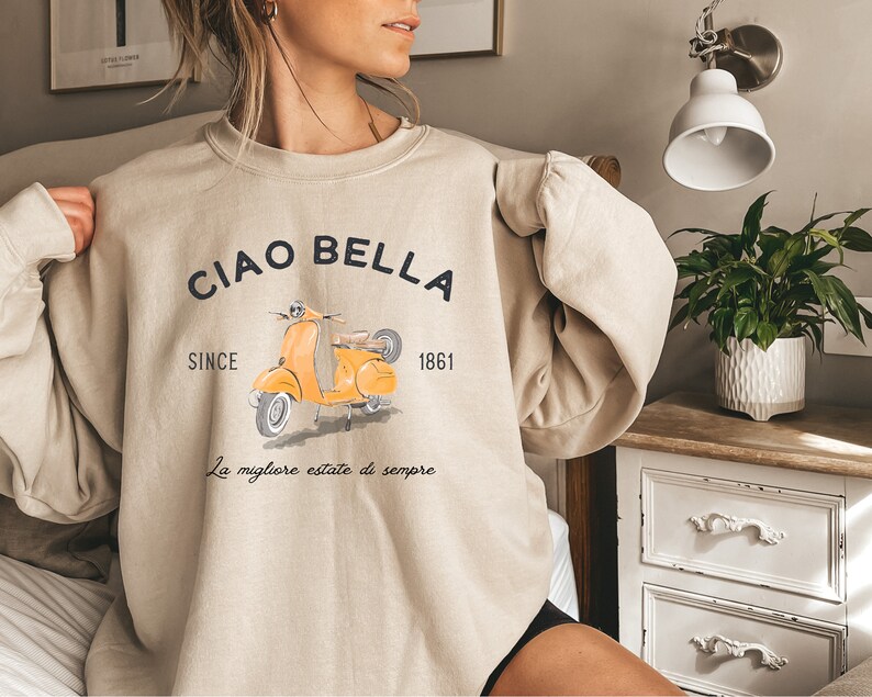 K&ouml;nnte beinhalten: Beigefarbener Pullover mit einem Aufdruck eines gelben Rollers und dem Text "Ciao Bella Since 1861 La migliore estate di sempre".