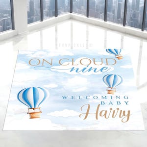 May include: A baby shower welcome sign with a blue and white hot air balloon theme. The sign reads "On Cloud Nine" and "Welcoming Baby Harry". The background is a watercolor sky with clouds.