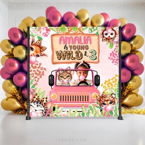 May include: A pink and white birthday party backdrop with the text "AMALIA is YOUNG WILD & 3". The backdrop features a pink jeep with animal characters and a child blowing a bubble. Balloons in gold and magenta surround the backdrop.