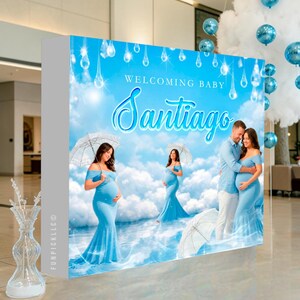 May include: A baby shower backdrop with a blue sky theme. The backdrop features the text "Welcoming Baby Santiago" and images of a pregnant person in a blue dress, a couple, and umbrellas. Blue balloons and cloud decorations are in the background.