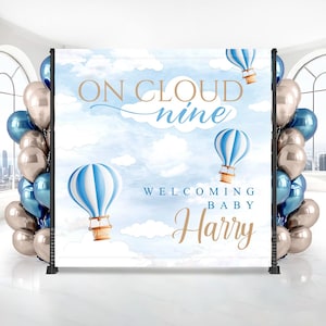 May include: A baby shower backdrop with a blue sky and cloud design, featuring hot air balloons. The text reads "ON CLOUD nine WELCOMING BABY Harry". Balloons in shades of blue and silver frame the backdrop.