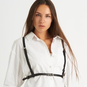 May include: A black leather harness with adjustable straps and a buckle closure. The harness is worn over a white button-down shirt.