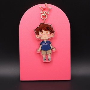 Puede incluir: Un llavero de dibujos animados con un personaje de pelo castaño, camisa azul y pantalones cortos beige. El llavero está sujeto a un cierre en forma de estrella. El fondo es un arco rosa sobre una superficie negra.