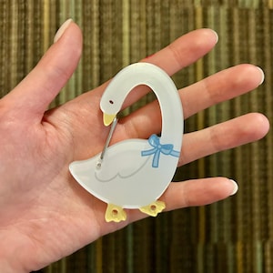 May include: A white swan-shaped brooch with a light blue bow and yellow feet. The brooch has a pin on the back for easy attachment. The swan's head is curved, and the design is simple and elegant.