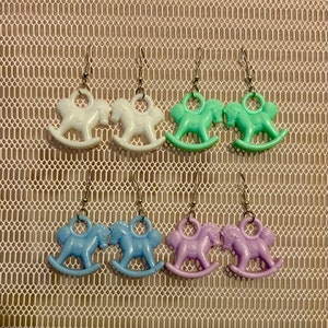 May include: Six pairs of rocking horse earrings in various pastel colors: white, mint green, light blue, and lavender. Each earring features a small, detailed rocking horse charm suspended from a silver-colored hook. The earrings are displayed against a textured, off-white background.