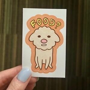 May include: A sticker featuring a cartoon dog with the word "FOOD?" above it. The dog is light beige with a pink nose and brown eyes. The text and dog are outlined in brown, with an orange border.