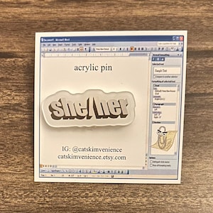May include: An acrylic pin with the text "she/her" in a bold, sans-serif font. The pin is set against a white background with a mock-up of a Microsoft Word document. The words "acrylic pin" are above the pin.