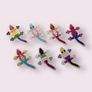 May include: A collection of seven beaded lizard pins in various colors. Each lizard is made of small, colorful beads, with different color combinations. The pins are arranged on a light pink background.