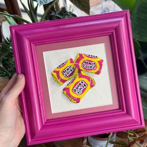 Könnte beinhalten: Eine gerahmte Stickerei von drei Fruit Salad Kaugummis, einer beliebten britischen Süßigkeit. Die Stickerei ist in rosa, gelben und weißen Fäden auf einem cremefarbenen Stoff ausgeführt. Der Rahmen ist leuchtend pink.