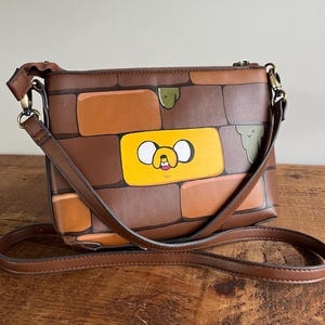 Può includere: Borsa a tracolla marrone con motivo a mattoni. La borsa presenta un rettangolo giallo con la faccia di un personaggio dei cartoni animati. La borsa ha una tracolla regolabile marrone e finiture color oro.