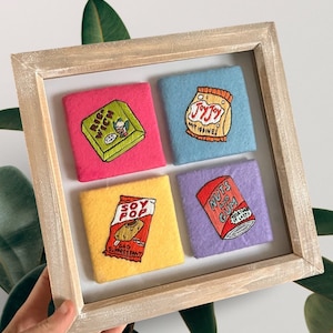 Può includere: Un'opera d'arte incorniciata con quattro quadrati di feltro ricamati. I quadrati, in rosa, azzurro, giallo e viola, raffigurano alimenti. I disegni includono un panino, un sacchetto di snack e una lattina di noci e gomma.