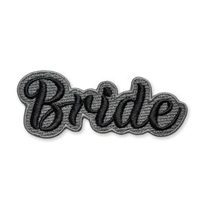 May include: A black embroidered patch with the word "Bride" in a cursive font. The patch has a gray border and is set against a white background. Ideal for bridal accessories.