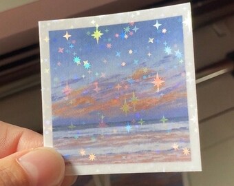 May include: A square sticker with a holographic finish featuring a sunset over a beach with a blue sky and stars. The sticker has a white border.