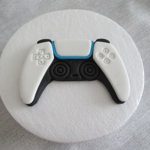 May include: A white and black video game controller cake topper with blue accents. The controller has a white top and black sides with a blue line across the top. The controller has four buttons on each side.