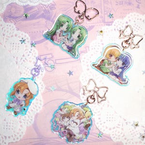 May include: Several anime-style acrylic charms featuring colorful illustrations of characters. Each charm has a different design, with images of pairs of characters hugging or posing. The charms are attached to metal keychains with bow or star accents.