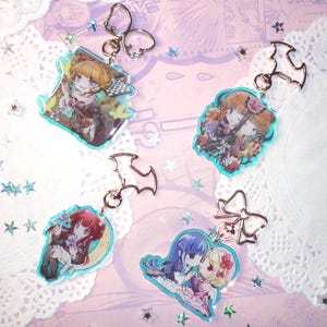 Può includere: Quattro portachiavi di personaggi in stile anime con bordi turchese e vari attacchi a ciondolo. I portachiavi presentano illustrazioni colorate di ragazze in abiti dettagliati. Lo sfondo è rosa chiaro con coriandoli a forma di stella e centrini di pizzo.