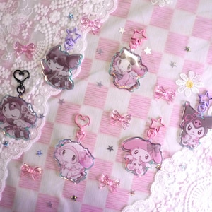 Peut inclure: Plusieurs porte-clés roses et violets avec des personnages de dessins animés, ornés d'attaches en forme d'étoile et de cœur. Ils sont disposés sur un fond à carreaux roses et blancs avec des nœuds et de la dentelle décoratifs.