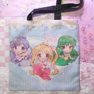 Puede incluir: Un bolso tote azul claro con un asa negra. La bolsa presenta tres personajes de sirenas de dibujos animados con cabello morado, rubio y verde. La bolsa tiene un fondo azul claro con un ribete de encaje blanco.