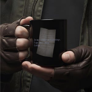 Peut inclure: Une tasse à café noire avec un motif de note manuscrite. Le texte sur la tasse se lit comme suit : "In my restless mornings, I see that mug. Coffee." La tasse est tenue par une personne portant des gants en cuir marron.