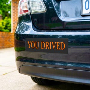 Puede incluir: Una pegatina rectangular negra con las palabras "YOU DRIVED" en letras naranjas está adherida a la parte trasera de un coche azul oscuro. El piloto trasero y la matrícula del coche son visibles.