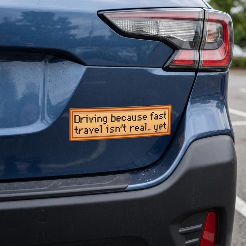 May include: A rectangular bumper sticker with the text "Driving because fast travel isn't real.. yet" in a pixelated font. The sticker is orange with a brown border and is affixed to the rear of a blue car.