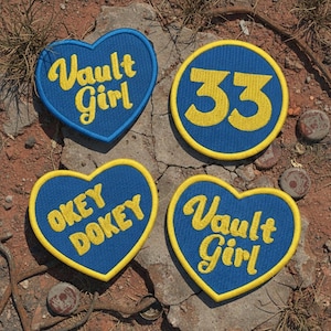 May include: Four embroidered patches. Two are heart-shaped, with the words "Vault Girl" and "Okey Dokey" in yellow script on a blue background. The other two are circular, one with the number "33" in yellow on a blue background.