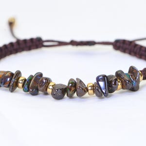 May include: A brown macrame bracelet featuring iridescent, dark-colored stone beads and small gold-colored beads. The bracelet has an adjustable sliding knot closure. The stones have a mix of brown, blue, and green hues.