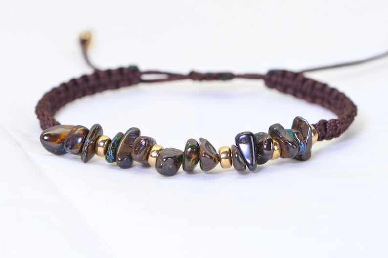 May include: A brown macrame bracelet featuring iridescent, dark-colored stone beads and small gold-colored beads. The bracelet has an adjustable sliding knot closure. The stones have a mix of brown, blue, and green hues.