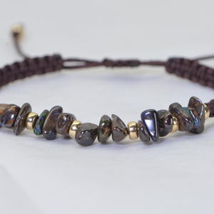 May include: A brown macrame bracelet featuring dark, iridescent gemstone chips and small gold-colored beads. The adjustable bracelet has a sliding knot closure. The gemstones have shades of brown, black, and hints of blue and green.
