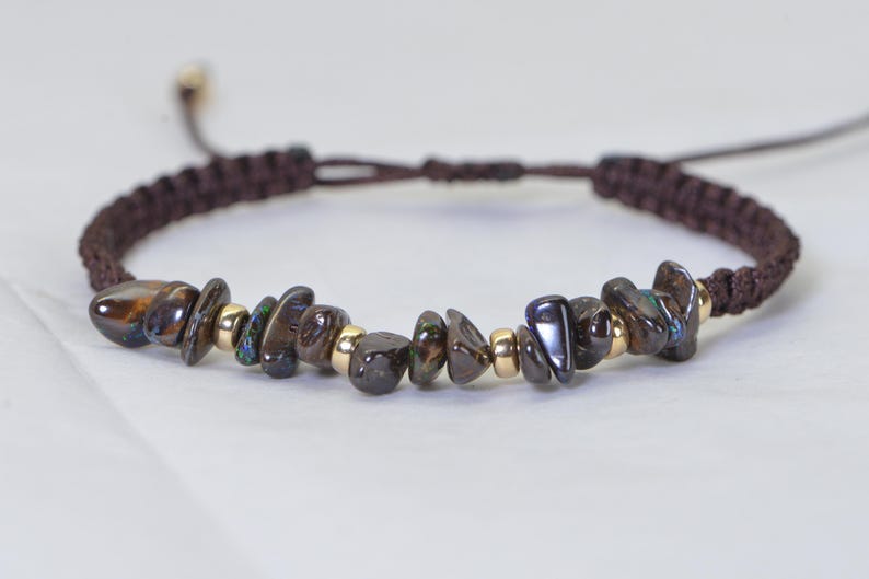 May include: A brown macrame bracelet featuring dark, iridescent gemstone chips and small gold-colored beads. The adjustable bracelet has a sliding knot closure. The gemstones have shades of brown, black, and hints of blue and green.