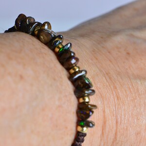 May include: A dark brown beaded bracelet with gold-colored accents. The bracelet features irregularly shaped, dark brown beads with hints of green and purple, strung together with gold-colored beads and brown cord.