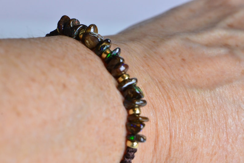 May include: A dark brown beaded bracelet with gold-colored accents. The bracelet features irregularly shaped, dark brown beads with hints of green and purple, strung together with gold-colored beads and brown cord.