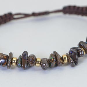May include: A brown macrame bracelet featuring a row of polished, dark brown and iridescent gemstone chips interspersed with small gold-colored beads. The bracelet has a sliding knot closure for adjustable sizing.