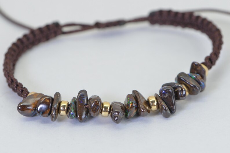 May include: A brown macrame bracelet featuring a row of polished, dark brown and iridescent gemstone chips interspersed with small gold-colored beads. The bracelet has a sliding knot closure for adjustable sizing.