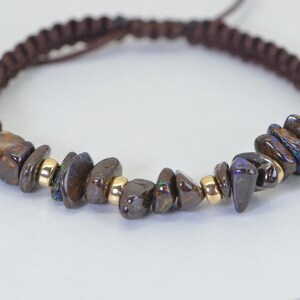 May include: A brown macrame bracelet featuring polished, dark brown opal stones with iridescent blue and green flashes. The bracelet includes small gold-colored beads and a sliding knot closure.
