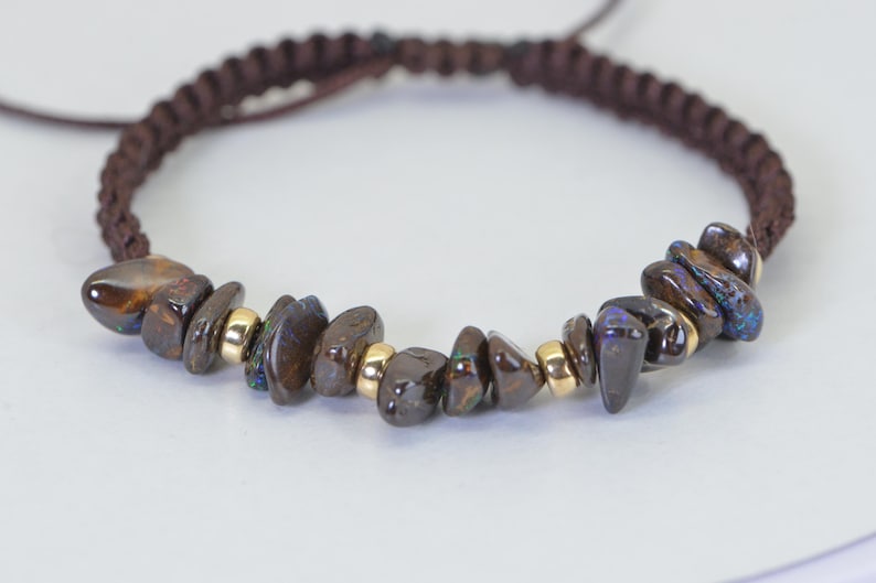 May include: A brown macrame bracelet featuring polished, dark brown opal stones with iridescent blue and green flashes. The bracelet includes small gold-colored beads and a sliding knot closure.