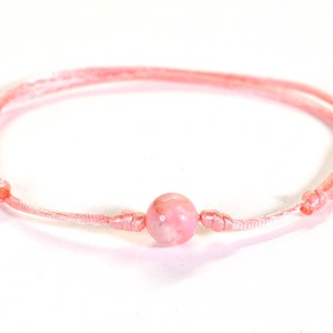 Puede incluir: Un brazalete de cuerda rosa con una sola cuenta de piedra preciosa rosa.