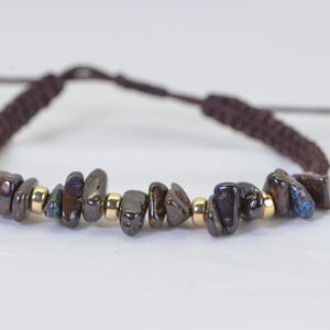 May include: A handmade bracelet featuring dark, irregularly shaped stones in shades of brown, black, and blue, interspersed with small gold-colored beads. The bracelet is strung on a brown, braided cord, with adjustable sliding knots.