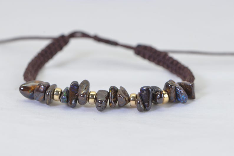 May include: A handmade bracelet featuring dark, irregularly shaped stones in shades of brown, black, and blue, interspersed with small gold-colored beads. The bracelet is strung on a brown, braided cord, with adjustable sliding knots.