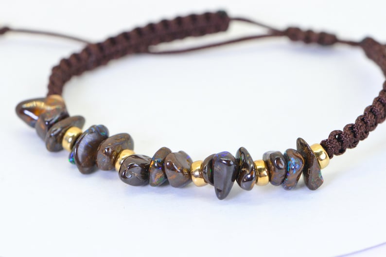 May include: A brown macrame bracelet featuring dark brown, irregularly shaped stones interspersed with small gold-colored beads. The bracelet has an adjustable closure.
