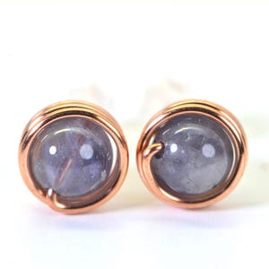 Peut inclure: Une paire de boucles d'oreilles à clous avec des pierres rondes, violet clair, enveloppées dans du fil de cuivre. Le fil de cuivre est enroulé autour de la pierre, créant un cadre décoratif. Fond blanc.
