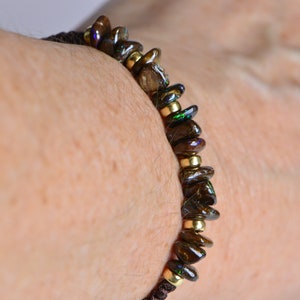 May include: A beaded bracelet featuring dark brown, iridescent stones and small gold-colored beads. The bracelet is strung on a dark brown cord. The bracelet is wrapped around a person's wrist.