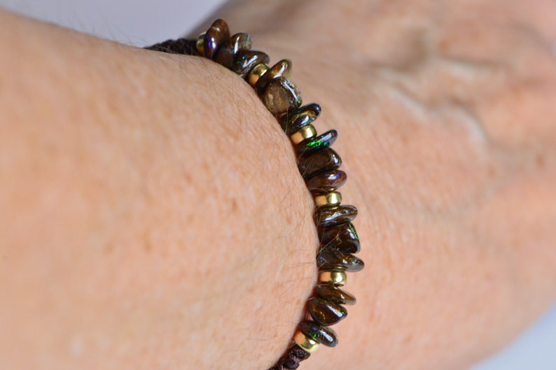 May include: A beaded bracelet featuring dark brown, iridescent stones and small gold-colored beads. The bracelet is strung on a dark brown cord. The bracelet is wrapped around a person's wrist.