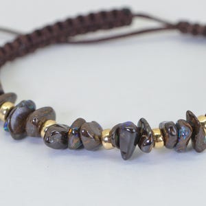 May include: Brown braided bracelet with dark brown, irregularly shaped stones and small gold-colored beads. The bracelet has an adjustable sliding knot closure. The stones have iridescent blue and green flashes.