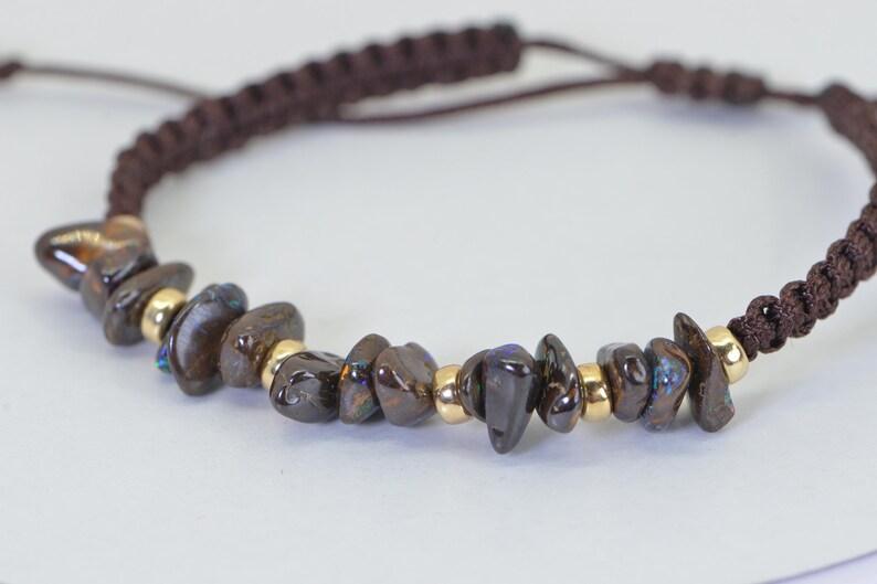 May include: Brown braided bracelet with dark brown, irregularly shaped stones and small gold-colored beads. The bracelet has an adjustable sliding knot closure. The stones have iridescent blue and green flashes.