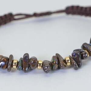 May include: A brown macrame bracelet featuring a row of dark, iridescent gemstone chips interspersed with small gold beads. The bracelet has a braided cord closure.