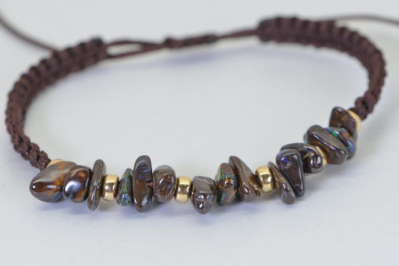 May include: A brown macrame bracelet featuring a row of dark, iridescent gemstone chips interspersed with small gold beads. The bracelet has a braided cord closure.
