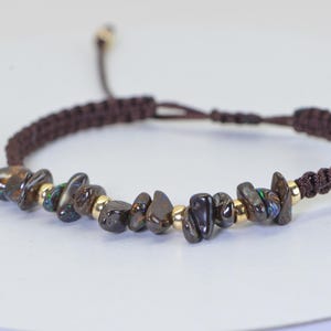 May include: Brown macrame bracelet with dark, irregularly shaped stones and small gold-colored beads. The bracelet has an adjustable sliding knot closure. The stones have iridescent green and blue flashes.