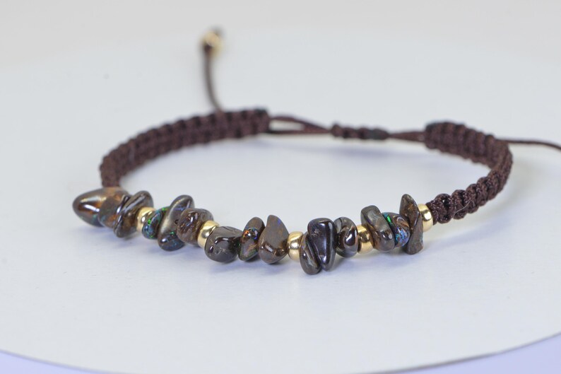 May include: Brown macrame bracelet with dark, irregularly shaped stones and small gold-colored beads. The bracelet has an adjustable sliding knot closure. The stones have iridescent green and blue flashes.